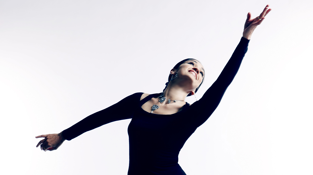 A female dancer in black clothes smiles and stretches her hand towards the sky